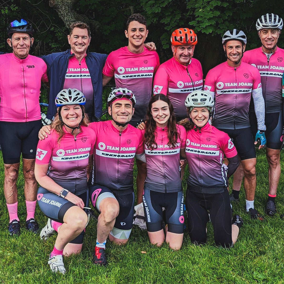 A team of cyclists posing together for a photo. There are two rows of people, three women and a man kneeling in the front, and six men standing in the back. The whole team is wearing custom pink and black cycling kits made by KindHuman Bicycles.